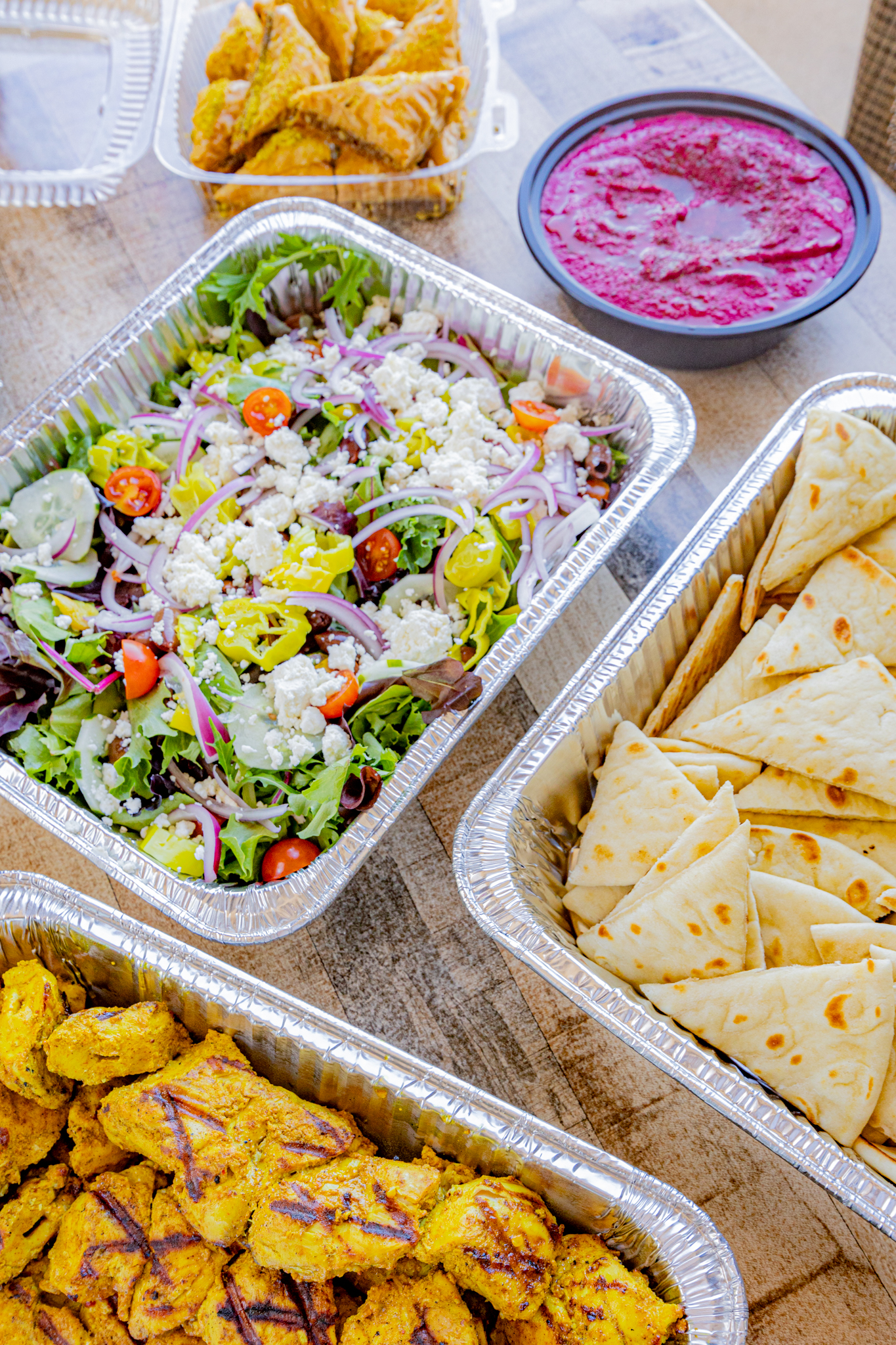 Portara catering trays filled with Greek salad, pita, chicken kabob, beet hummus, and baklava.