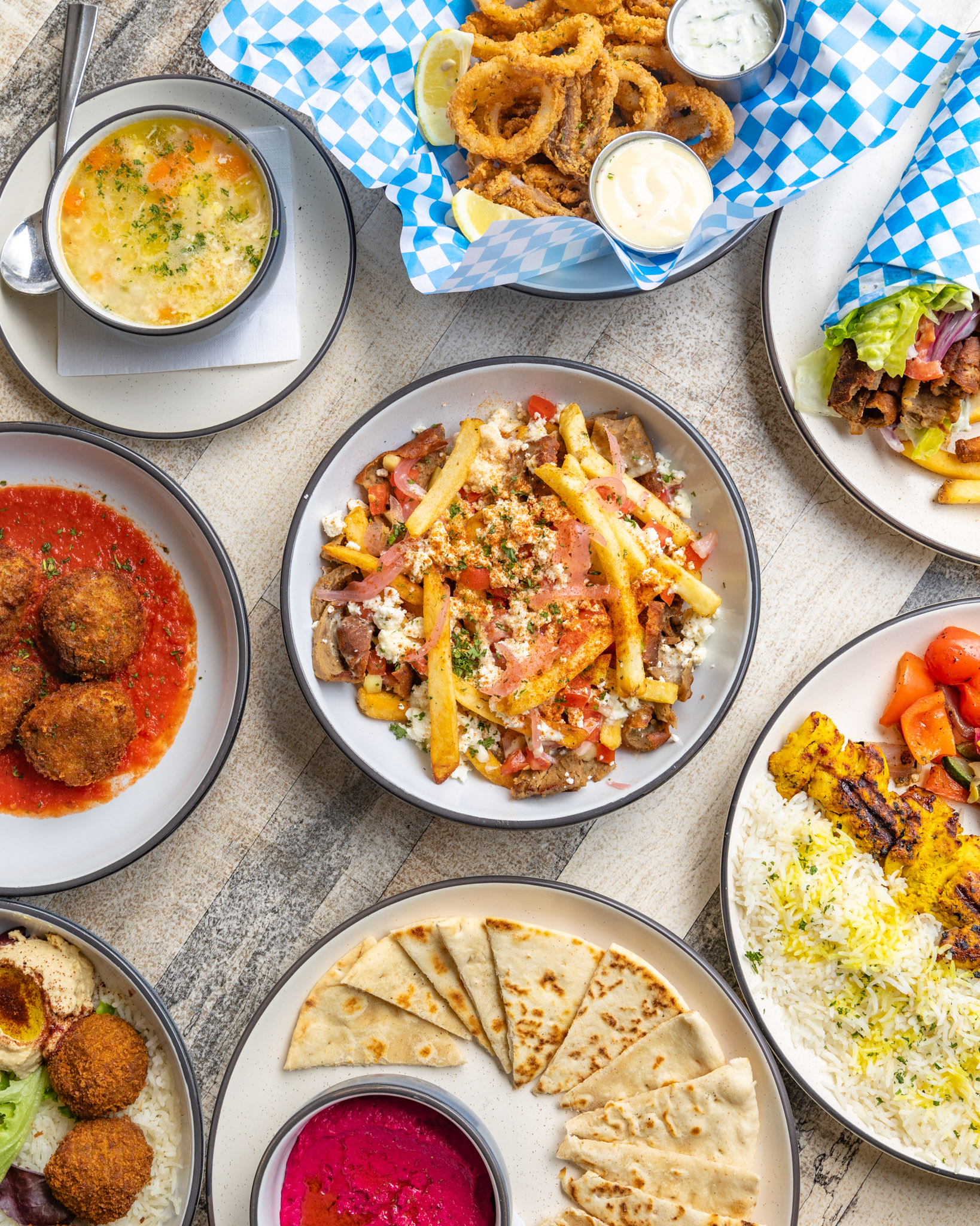 A colorful table filled with Portara soup, calamari, loaded fries, pita, rice, kabobs, and hummus.