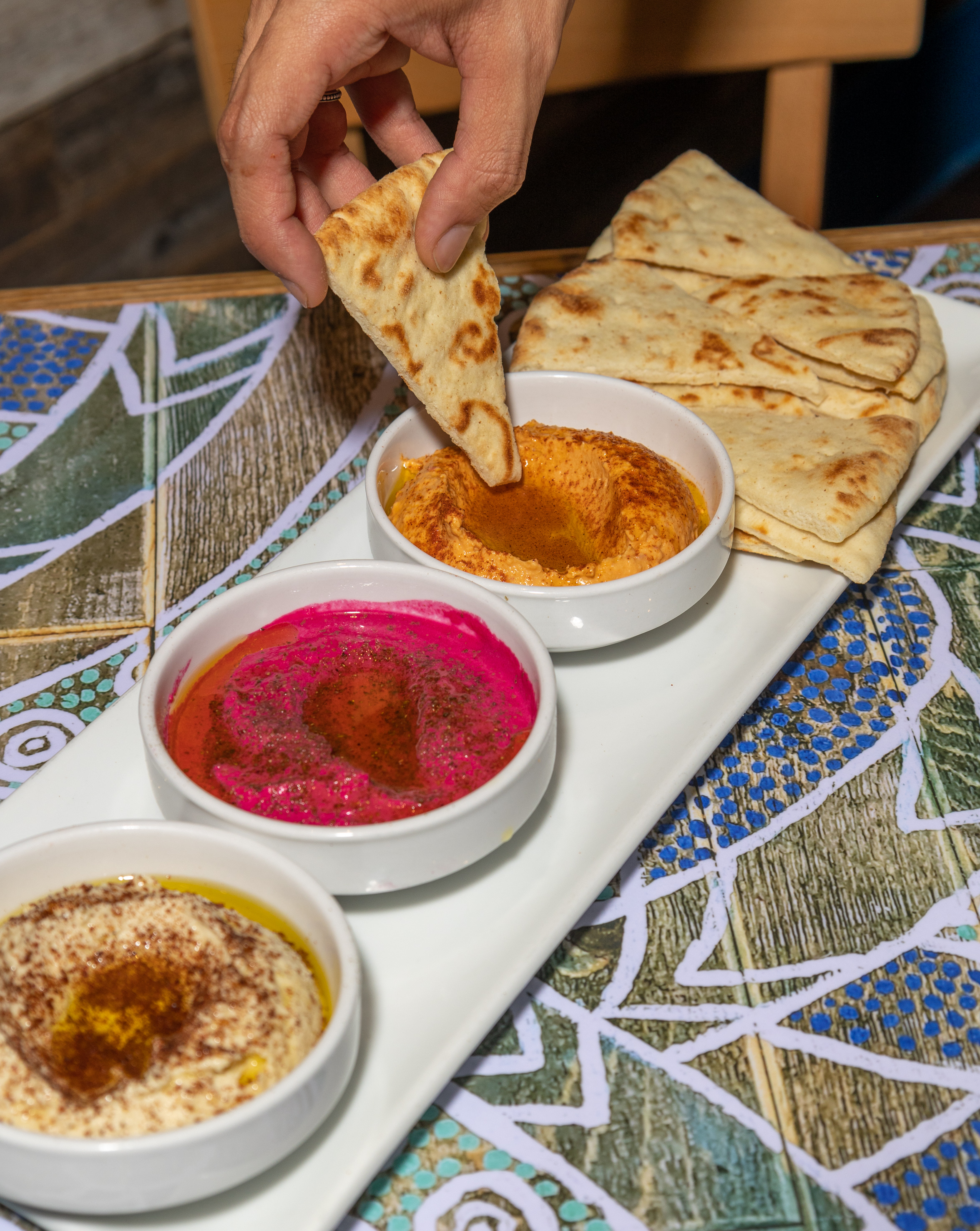 A guest dipping warm pita into a colorful spread of Portara hummus.