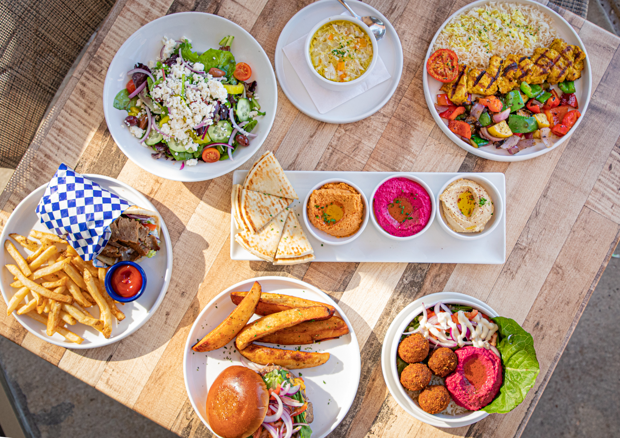 A colorful spread of Portara salads, hummus, pita, grilled meats, fries, and bowls.